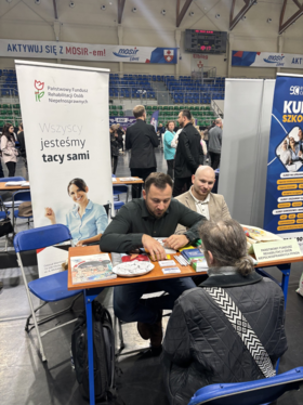 Stoisko informacyjne w hali. Przy stoliku siedzą osoby, a obok znajduje się stojący baner z napisem „Wszyscy jesteśmy tacy sami”. W tle widać kolejne osoby oraz stoiska wystawiennicze. Na stoliku leżą materiały informacyjne.