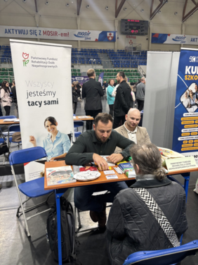 Pokaż zdjęcie: Stoisko informacyjne w hali. Przy stoliku siedzą osoby, a obok znajduje się stojący baner z napisem „Wszyscy jesteśmy tacy sami”. W tle widać kolejne osoby oraz stoiska wystawiennicze. Na stoliku leżą materiały informacyjne.