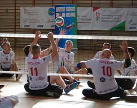 Pokaż zdjęcie: Zawodnicy ze Szczecina siedzący na boisku przekazują przez siatkę piłkę między drużynami. W tle widać drabinki i sportowe i loga sponsorów, w tym PFRON