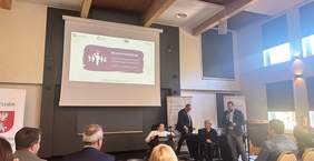 Pokaż zdjęcie: W sali konferencyjnej odbywa się spotkanie. Na ścianie wisi ekran projekcyjny z prezentacją zatytułowaną Własnym Głosem – 9 Podlaski Konwent Osób z Niepełnosprawnościami Augustów 18-19 października 2024 r. Na pierwszym planie widoczni są prelegenci, w tym dwie osoby na wózkach inwalidzkich.  Jeden z mężczyzn stoi przy mikrofonie, drugi stoi za kobietami siedzącymi na wózkach.