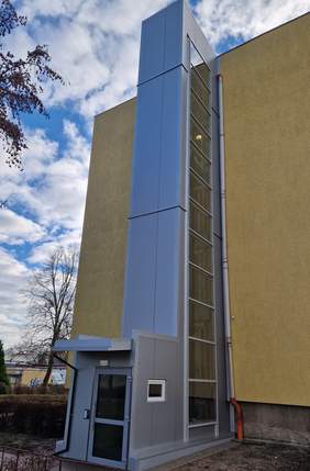 Pokaż zdjęcie: Zewnętrzna winda dobudowana do wielokondygnacyjnego budynku mieszkalnego. Konstrukcja windy jest nowoczesna, w kolorze szarym, z przeszklonym szybem. Winda stoi przy żółtej elewacji bloku, a wokół znajdują się chodnik, krzewy i drzewo bez liści.