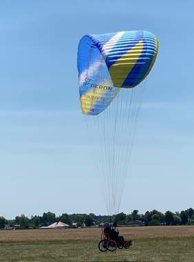 Pokaż zdjęcie: Uczestnik szkolenia na wózku inwalidzkim, a do niego przypięta paralotnia nabiera prędkości  w celu oderwania się od ziemi.