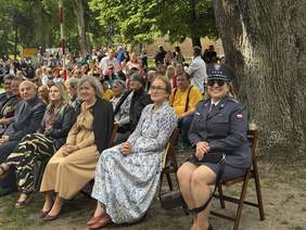 Pokaż zdjęcie: Goście zaproszeni na festiwal siedzą na ławeczkach ustawionych na trawie.