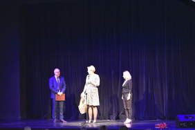 Pokaż zdjęcie: Na scenie stoi mężczyzna oraz dwie kobiety, jedna z nich trzyma w ręku kosz z cukierkami oraz przemawia do mikrofonu