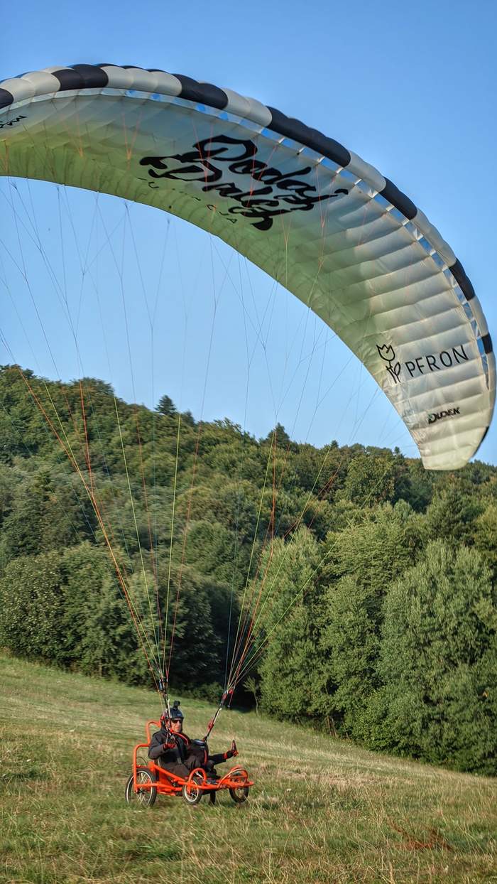 Pokaż zdjęcie: Na trawiastym zboczu ląduje mężczyzna lecący paralotnią oznaczoną logotypami PFRON oraz Fundacji im. Doktora Piotra Janaszka Podaj Dalej. W tle widoczny jest las oraz bezchmurne niebo.