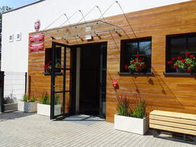 Pokaż zdjęcie: Zdjęcie wejścia do budynku, na parapetach stoją doniczki z czerwonymi pelargoniami, przy drzwiach wejściowych stoi ławeczka. Elewacja w kolorze drewna.