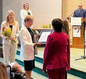 Pokaż zdjęcie: Na scenie podczas gali Dyrektor Oddziału Podlaskiego PFRON Edyta Kondzior w białej marynarce wręcza dyplom i statuetkę LODOŁAMACZ 2025 przedstawicielkom Fundacji Hospicjum Proroka Eliasza. Obok stoją uczestnicy uroczystości.