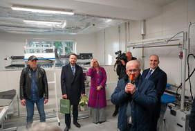 Grupa pięciu mężczyzn i jednej kobiety w jasnym, profesjonalnym pomieszczeniu z białymi, kafelkowymi ścianami. W tle widoczna jest maszyna przemysłowa i okno. Mężczyzna na pierwszym planie po prawej stronie mówi do mikrofonu. Jeden z mężczyzn w głębi trzyma kamerę.