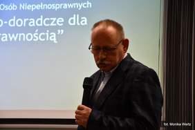 Pokaż zdjęcie: Prezes Stowarzyszenia na Rzecz Edukacji "Pomost" na tle wyświetlonej prezentacji, przemawia i trzyma w ręku mikrofon