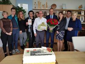 Grupa dziesięciu osób, kobiety i mężczyźni stojąc w szeregu pozują do zdjęcia. Są uśmiechnięci. Sytuacja ma miejsce w pokoju, stoją na tle mebli. Przed nimi na stole leży jasny, prostokątny tort przyozdobiony kwiatami i napisem