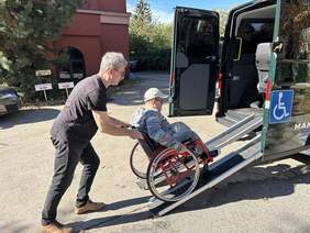 Pokaż zdjęcie: Na parkingu stoi samochód przystosowany do przewozu osób z niepełnosprawnością. Tylne drzwi pojazdu są otwarte. Do wnętrza auta wjeżdża osoba na wózku inwalidzkim. Wprowadza ją inny mężczyzna. 