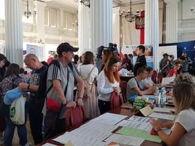 Duża sala, na niej targi pracy, część osób siedzi za stołami, pozostali chodzą, widoczne banery instytucji i pracodawców