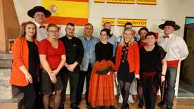 Pokaż zdjęcie: Na zdjęciu grupa uśmiechniętych ludzi. Część z nich ubrana w stylu hiszpańskim. Dwaj mężczyźni w białych koszulach z czerwonymi muszkami. Czarnych hiszpańskich kapeluszach przepasanych czerwonymi pasami. Kobieta po środku w czarnej bluzce i p