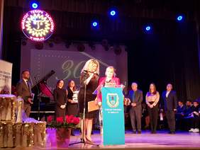 Na scenie z przodu dwie panie przemawiają, za nimi w szeregu stoi grupa kilku osób, mężczyzn i kobiet, całkiem na prawo widać siedzących uczestników.