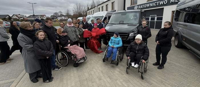 Pokaż zdjęcie: Grupa ludzi stoi przed budynkiem ratownictwa medycznego, obok czarnego busa. 