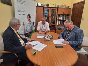 Pokaż zdjęcie: Przy owalnym stole siedzi trzech mężczyzn. Podpisują umowę. Na stole leżą różne dokumenty, pieczątki, telefony, długopisy. 