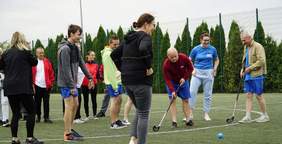 Pokaż zdjęcie: Grupa kilkunastu osób znajduje się na boisku ogrodzonym wysokim płotem. Dwóch mężczyzn trzyma w ręce kije do unihokeja. Na boisku znajduje się również mała, niebieska piłka do unihokeja. W tle żywopłot z tui.