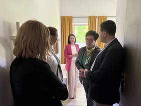 Pokaż zdjęcie: Grupa osób stoi w pokoju z żółtymi zasłonami, prowadzą rozmowę. Kobieta z kolorową apaszką mówi, reszta słucha. Pomieszczenie o domowym charakterze.