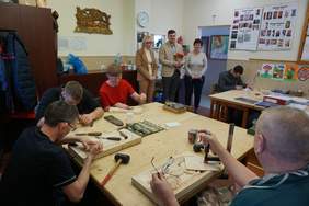 Pokaż zdjęcie: Zastajemy uczestników przy pracy przy płaskorzeźbach. Widać już zarysy słoneczników, Matki Boskiej oraz serca na dłoni. Na ścianie wisi dzieło skończone: płaskorzeźba ukazująca woźnice prowadzącego konia z wozem, których okalają drzewa