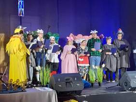 Pokaż zdjęcie: Na scenie występuje grupa teatralna. Aktorzy są przebrani za różne zwirzęta. Trzymają w dłoniach mikrofony, do których śpiewają. 