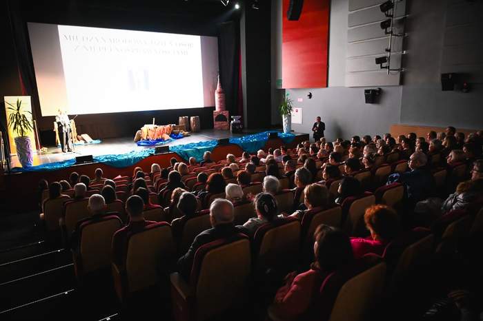 Pokaż zdjęcie: Szeroki plan widowni teatru lub sali kinowej, wypełnionej ludźmi. Na scenie po lewej stronie widać dekoracje imitujące pejzaż wodny i ruiny oraz postacie w strojach scenicznych. Na dużym ekranie w tle wyświetlony jest napis w języku polskim: "MIĘDZYNARODOWY DZIEŃ OSÓB Z NIEPEŁNOSPRAWNOŚCIAMI". Po prawej stronie sceny stoi mężczyzna w garniturze.