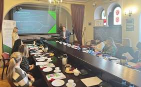 Pokaż zdjęcie: Sala konferencyjna z długim drewnianym stołem i ludźmi siedzącymi po obu jego stronach. Na środku stołu leżą liczne dokumenty, notesy i kolorowe broszury, stoją butelki wody, termosy z napojami, szklanki i małe talerzyki. Przy miejscach uczestników spotkania znajdują się mikrofony konferencyjne. Po obu stronach stołu siedzą osoby w eleganckich strojach, pochylone nad materiałami lub zwrócone w stronę przodu sali.   Przy przednim końcu stołu stoi kobieta w jasnym żakiecie, pochylona nad laptopem i prowadzi prezentację. Obok niej, z prawej strony przy ekranie projekcyjnym, stoi mężczyzna w ciemnej marynarce i jasnej koszuli. Na ścianie wyświetlona jest prezentacja w odcieniach zieleni i błękitu z dużym napisem „Środki PFRON” oraz słowami „oferta” i „cel”, a po prawej stronie widnieje logo Państwowego Funduszu Rehabilitacji Osób Niepełnosprawnych.