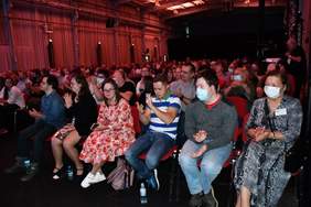 Pokaż zdjęcie: Fotografia przedstawia widownię. Widownia oświetlona jest czerwonym światłem, osoby siedzą na krzesłach i biją brawo dla występującego na scenie zespołu muzycznego.