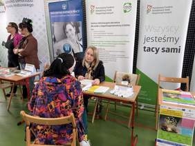 Pokaż zdjęcie: Stoisko informacyjne CIDON. Przy stole siedzi kobieta i mężczyzna, przed nimi ulotki i materiały. W tle banery, napis „Wszyscy jesteśmy tacy sami” oraz informacje o wsparciu dla osób z niepełnosprawnościami.