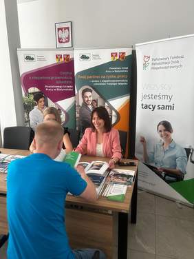 Pokaż zdjęcie: Na zdjęciu dwie kobiety i jeden mężczyzna siedzą przy stole, rozmawiając z kobietą siedzącą naprzeciwko. W tle widoczne są banery Państwowego Urzędu Pracy w Białymstoku i Państwowego Funduszu Rehabilitacji Osób Niepełnosprawnych. Na stole leżą materiały informacyjne.