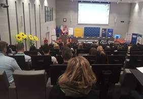 Pokaż zdjęcie: Scena konferencji z perspektywy górnych rzędów dużej sali. Z przodu sali, po lewej stronie znajduje się mównica, przy której stoi mężczyzna ubrany w marynarkę i krawat.