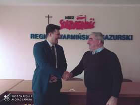 Pokaż zdjęcie: Dwóch uśmiechniętych mężczyzn podaje sobie dłoń, za nimi na ścianie napis ,,NSZZ Solidarność Region Warmińsko-Mazurski