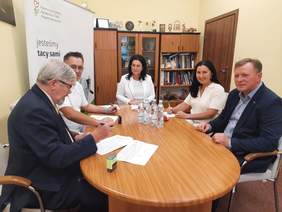 Pokaż zdjęcie: Przy owalnym stole siedzi pięć osób: dwie kobiety i trzech mężczyzn. Zawierają umowę. Na stole leżą różne dokumenty, pieczątki, woda mineralna. Niektórzy się uśmiechają. 