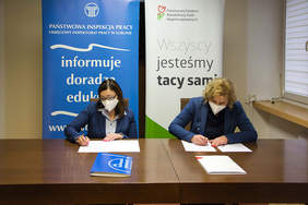 Pokaż zdjęcie: dwie kobiety w niebieskich marynarkach i maseczkach siedzą przy brązowym stole, przed nimi leżą dokumenty, które podpisują w tle loga PIP oraz PFRON