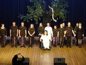 Pokaż zdjęcie: Na scenie znajdują się aktorzy. Kłaniają się po zakończonym występie. Jedna pani siedzi na wózku inwaidzkim. 