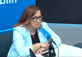 Pokaż zdjęcie: W studiu radiowym kobieta w niebieskiej marynarce i okularach udziela wywiadu.