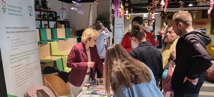 Pokaż zdjęcie: Grupa osób zebranych wokół stoiska informacyjnego. Po lewej stronie stoi roll-up Państwowego Funduszu Rehabilitacji Osób Niepełnosprawnych oraz niżej tekst: „Centra informacyjno-doradcze dla osób z niepełnosprawnościami” zwane CIDON. Przy stanowisku informacyjnym stoi pracownik funduszu i udziela informacji uczestnikom targów.