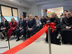 Pokaż zdjęcie: Zdjęcie na którym widać kawałek symbolicznej czerwonej szarfy, w tle na krzesełkach siedzą zaproszeni goście.