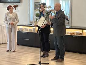 Pokaż zdjęcie: Przy mikrofonie stoi mężczyna w garniturze, obok dwie uśmiechnięte kobiety. Zdjęcie w sali restauracyjnej podczas części oficjalnej.