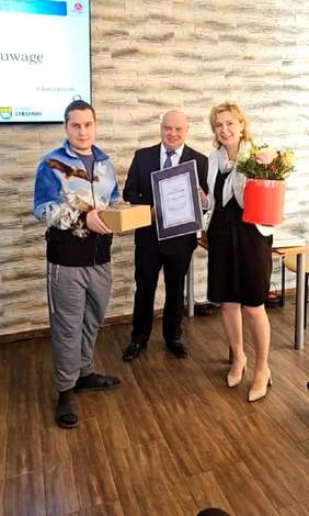 Pokaż zdjęcie: Na zdjęciu do fotografii pamiątkowej pozują trzy osoby, dwóch mężczyzn i kobieta stojąca po prawej stronie. Mężczyzna na środku ubrany jest w ciemny garnitur, trzyma w ręku dyplom, kobieta trzyma w ręku box z kwiatkami