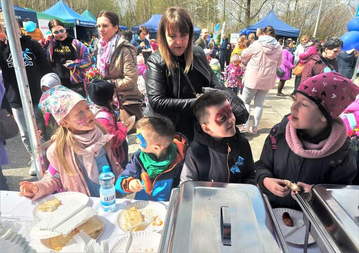 Pokaż zdjęcie: Zdjęcie festynu, dużej liczby osób przy stoiskach i kramach. Na pierwszym planie dzieci z twarzami pomalowanymi w motylki.