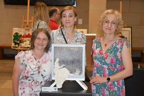 Pokaż zdjęcie: Trzy kobiety stoi przy pracy plastycznej, patrzą wprost w obiektyw