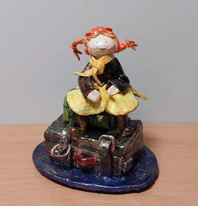 Pokaż zdjęcie: rzeźba przedstawia dziewczynkę z kucykami w żółtej sukience, siedzącą na kufrze