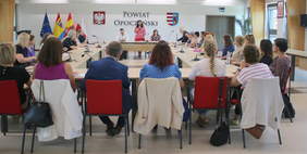 Pokaż zdjęcie: W sali konferencyjnej stoją stoły ustawione w kwadrat. Z każdej strony siedzą na krzesłach ludzie. Na stołach leżą mikrofony, dokumenty, notesy. 