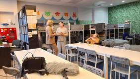 Pokaż zdjęcie: W ośrodku recepcyjnym w sali dziennej, gdzie stoją piętrowe łóżka rozmawiają dwie kobiety. Jedna z nich blondynka ubrana jest w beżowy garnitur, druga z pań ma na sobie dresy, jest mieszkanką ośrodka. Na stole leży mikron oraz futerał ze stacją nagłaśniającą.. 