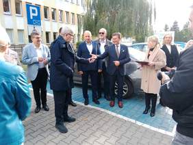 Pokaż zdjęcie: Jeden mężczyzna wręcza drugiemu kluczyki od samochodu. W tle widać także inne osoby. 