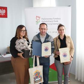 Trzy osoby stoją przed roll‑upem Państwowego Funduszu Rehabilitacji Osób Niepełnosprawnych, trzymając dyplomy i upominki, w tym torbę z kolorową grafiką.