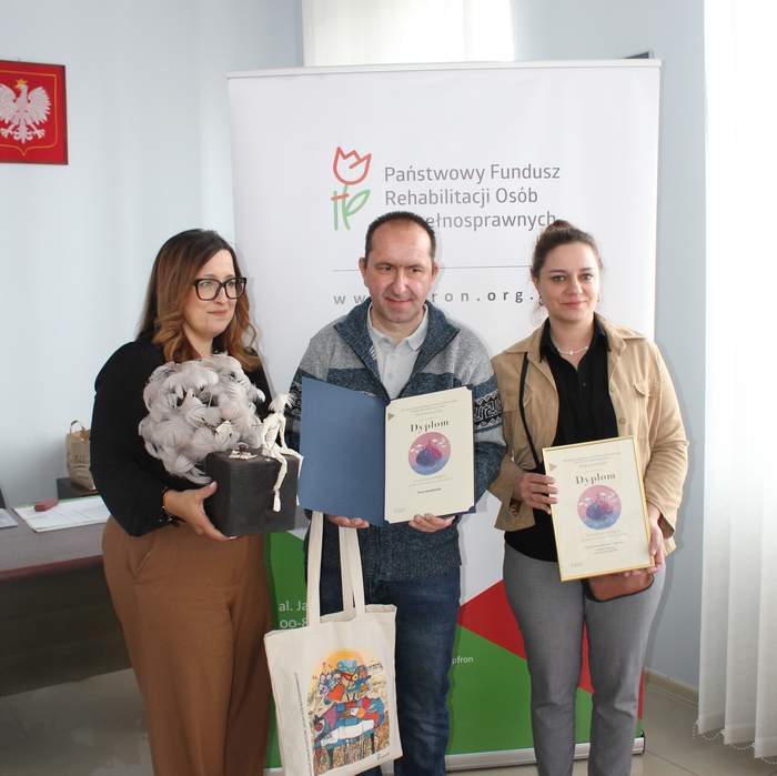 Pokaż zdjęcie: Trzy osoby stoją przed roll‑upem Państwowego Funduszu Rehabilitacji Osób Niepełnosprawnych, trzymając dyplomy i upominki, w tym torbę z kolorową grafiką.