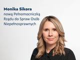 W prawej części grafiki widoczna górna część sylwetki kobiety. Kobieta ma jasne falowane włosy sięgające za ramiona. Ubrana w czarną bluzkę w białe kropki. Lekko uśmiecha się i patrzy w stronę aparatu. W lewej części grafiki tekst: Monika Sikora nową Pełnomocniczką Rządu do Spraw Osób Niepełnosprawnych. Całość na jasnoszarym tle.
