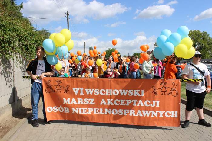 Pokaż zdjęcie: Uczestnicy Marszu w trakcie pochodu, trzymają baner w kolorze pomarańczowym z napisem "VI Marsz Akceptacji Osób Niepełnosprawnych"