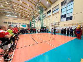 Zawodnicy siedzący na parkiecie rozgrywają mecz siatkówki na siedząco. Po obu stronach siatki widać drużyny w czerwonych i czarnych strojach sportowych, a w tle znajduje się zawodniczka przygotowująca się do zagrania piłki.